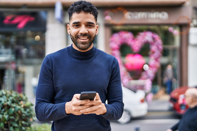 Joven hispano sonriendo confiado usando un teléfono inteligente en la calle