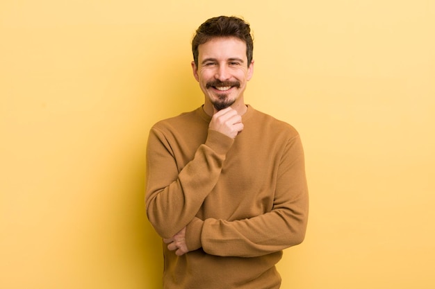 Joven hispano sonriendo disfrutando de la vida sintiéndose feliz amistoso satisfecho y despreocupado con la mano en la barbilla