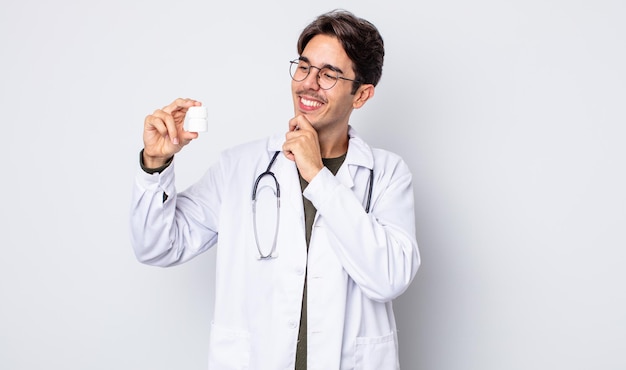 Joven hispano sonriendo con una expresión feliz y segura con la mano en la barbilla. médico con concepto de botella de píldoras