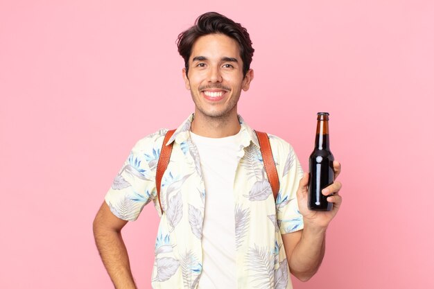 Joven hispano sonriendo felizmente con una mano en la cadera y confiado y sosteniendo una botella de cerveza