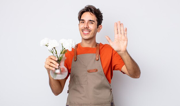 Joven hispano sonriendo felizmente, saludando con la mano, dándote la bienvenida y saludándote. concepto de floristería