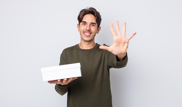 Joven hispano sonriendo y mirando amigable, mostrando el número cinco. concepto de caja blanca