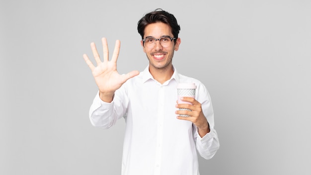 Joven hispano sonriendo y mirando amigable, mostrando el número cinco y sosteniendo un café para llevar