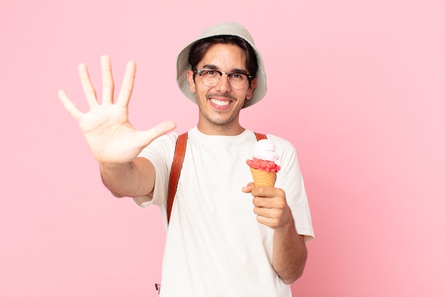 Joven hispano sonriendo y mirando amigable, mostrando el número cinco y sosteniendo un helado