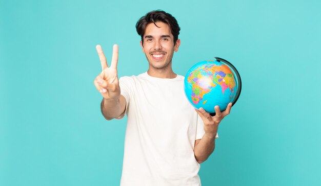 Joven hispano sonriendo y mirando amigable, mostrando el número dos y sosteniendo un mapa del globo terráqueo