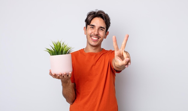 Joven hispano sonriendo y mirando feliz, gesticulando victoria o paz. concepto de planta decorativa