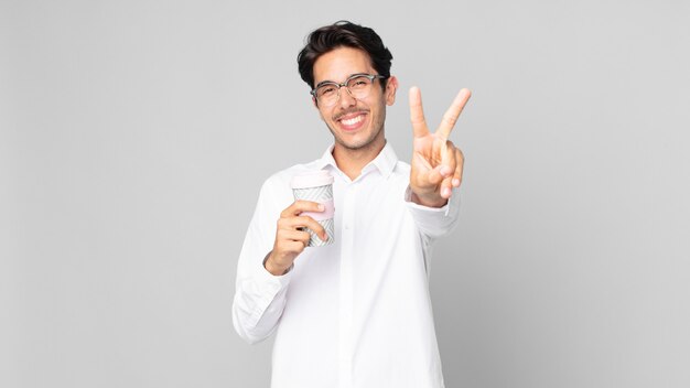 Joven hispano sonriendo y mirando feliz, gesticulando victoria o paz y sosteniendo un café para llevar