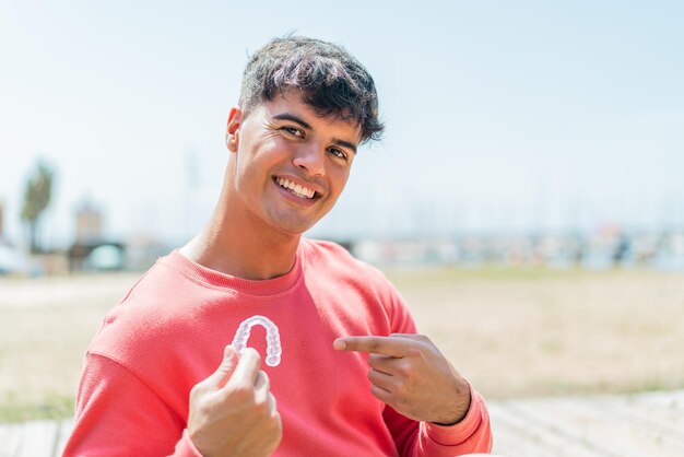 Foto joven hispano sosteniendo aparatos dentales invisibles al aire libre y apuntándolo