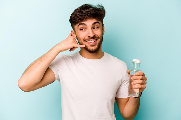 Joven hispano sosteniendo una botella de agua aislada de fondo blanco mostrando un gesto de llamada de teléfono móvil con los dedos