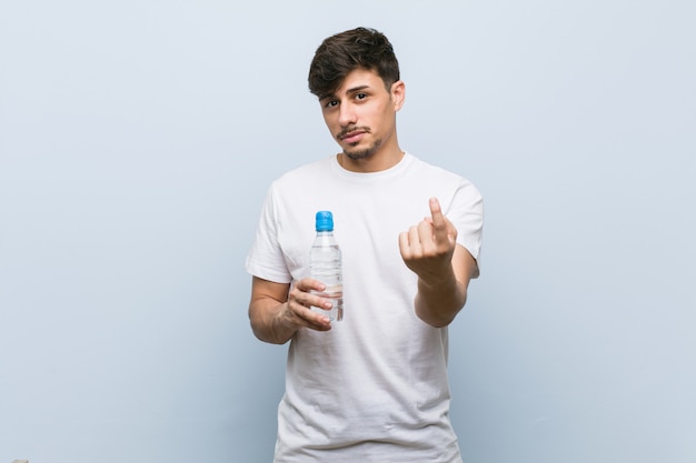 Joven hispano sosteniendo una botella de agua apuntando con el dedo hacia usted como invitando a acercarse.