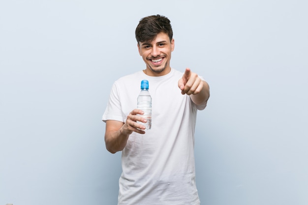 Joven hispano sosteniendo una botella de agua sonrisas alegres apuntando al frente.