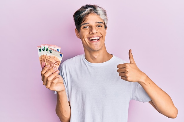 Joven hispano sosteniendo un montón de billetes de 10 euros sonriendo feliz y positivo, pulgar arriba haciendo excelente y señal de aprobación