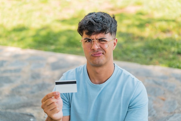 Joven hispano sosteniendo una tarjeta de crédito al aire libre con expresión triste