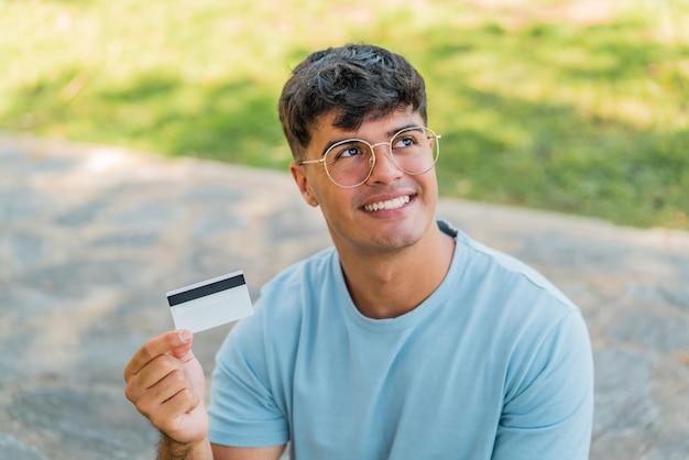 Joven hispano sosteniendo una tarjeta de crédito al aire libre mirando hacia arriba mientras sonríe