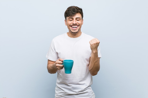 Joven hispano sosteniendo una taza animando despreocupado y emocionado. Concepto de victoria