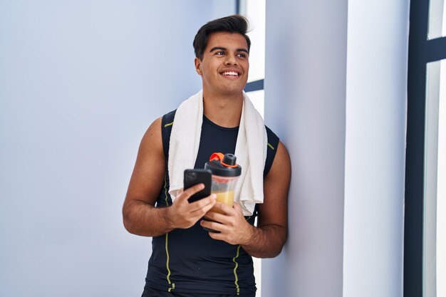 Joven hispano usando un teléfono inteligente bebiendo batido de proteínas en el centro deportivo