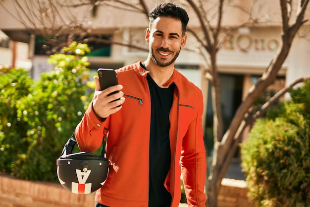 Joven hispano usando un teléfono inteligente con casco de moto en la ciudad.