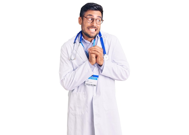 Joven hispano vestido con uniforme médico y estetoscopio riendo nervioso y emocionado con las manos en la barbilla mirando hacia un lado