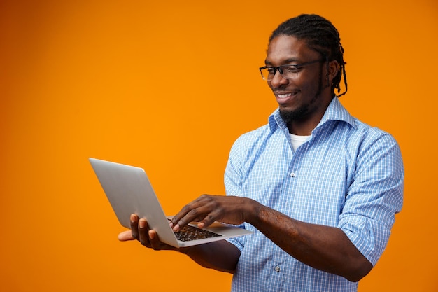 Joven hombre africano sonriente de pie y usando una computadora portátil sobre un fondo amarillo