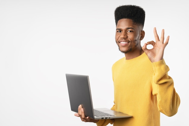 Joven hombre afroamericano trabajando usando una computadora portátil de pie sobre un fondo blanco haciendo OK con los dedos