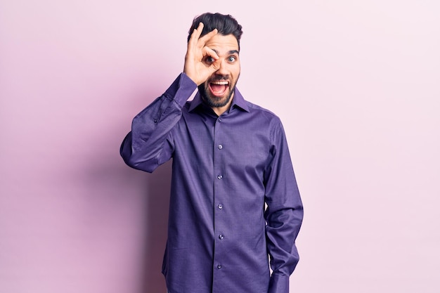 Joven hombre apuesto con barba y camisa informal haciendo un buen gesto con la mano sonriendo, mirando los ojos a través de los dedos con cara feliz.