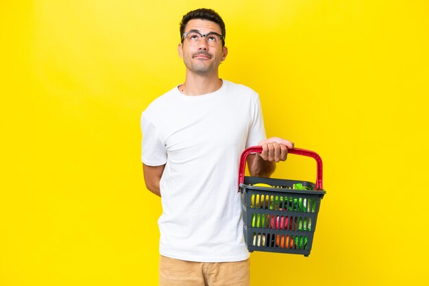 Joven hombre apuesto sosteniendo una cesta de la compra llena de comida sobre un fondo amarillo aislado y mirando hacia arriba