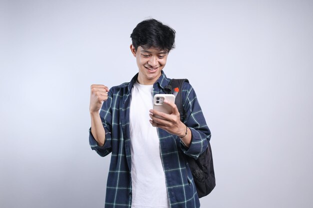 Joven hombre asiático caucásico con mochila mirando su teléfono inteligente con el puño cerrado mostrando el gesto de victoria