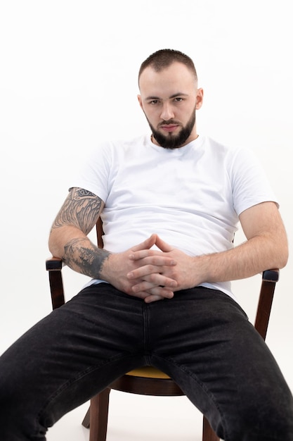 Joven hombre barbudo tranquilo con cabello oscuro corto en camiseta blanca jeans negros sentado en una silla de madera inte