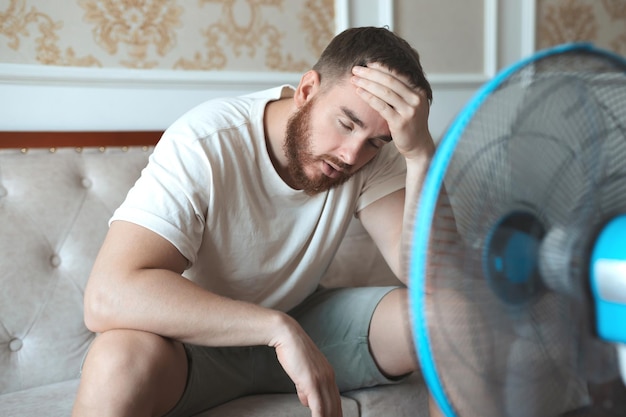 Joven hombre barbudo usando ventilador eléctrico en casa sentado en el sofá enfriándose durante el clima caluroso