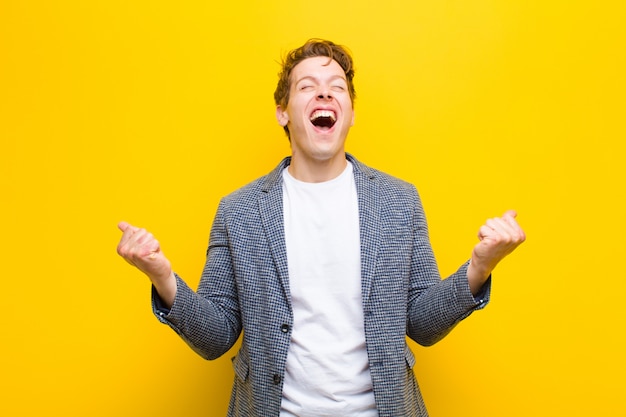 Joven hombre de cabeza roja que parece extremadamente feliz y sorprendido, celebrando el éxito, gritando y saltando sobre fondo naranja