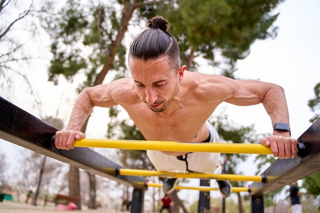 Joven hombre caucásico sin camisa haciendo flexiones en una barra en un parque de calistenia al aire libre