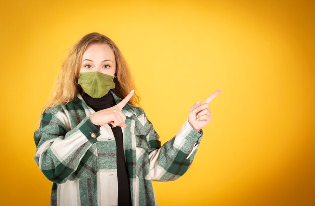 Joven hombre caucásico feliz apuntando con el dedo hacia arriba para copiar el espacio con una máscara higiénica para prevenir la infección por el virus corona sobre fondo amarillo