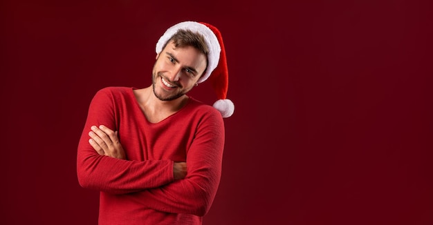 Joven hombre caucásico guapo en suéter rojo y sombreros de Santa está de pie sobre fondo rojo en el estudio y