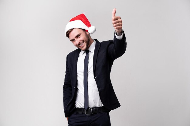 Joven hombre caucásico guapo en traje de negocios y sombreros de Santa se para sobre un fondo blanco en el estudio