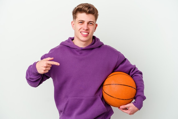 Joven hombre caucásico jugando baloncesto aislado fondo persona apuntando con la mano a un espacio de copia de camisa, orgulloso y seguro