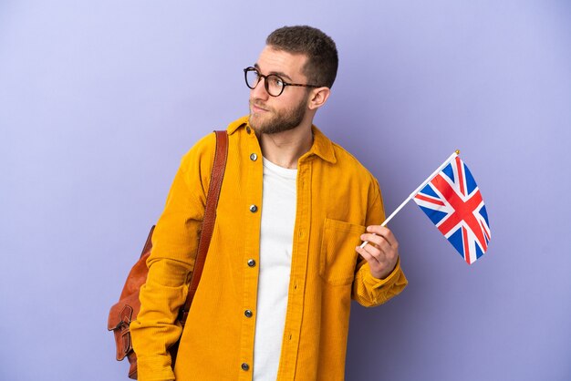 Joven hombre caucásico sosteniendo una bandera del Reino Unido aislado sobre fondo púrpura mirando hacia el lado