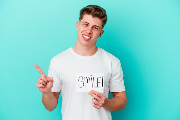 Joven hombre caucásico sosteniendo un cartel de sonrisa aislado sobre fondo azul.