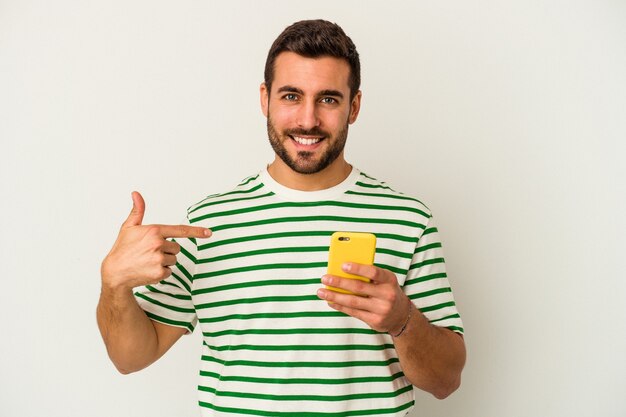 Joven hombre caucásico sosteniendo un teléfono móvil aislado sobre fondo blanco persona apuntando con la mano a una camisa, orgulloso y seguro