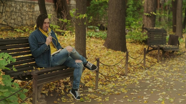 Joven hombre creativo con rastas escribiendo en un cuaderno sentado en un banco del parque en otoño dorado