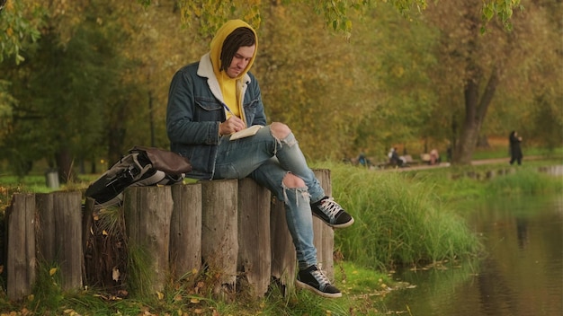 Joven hombre creativo con rastas escribiendo en un cuaderno sentado en tocones cerca del lago en un parque
