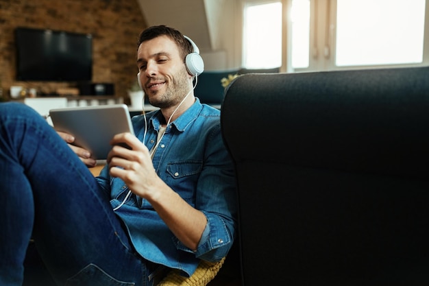 Joven hombre feliz con auriculares navegando por la red en una tableta digital mientras se relaja en la sala de estar.