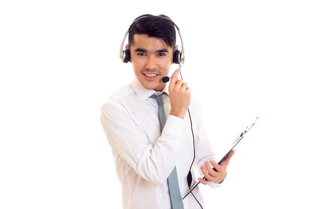 Joven hombre feliz con cabello negro en camisa blanca con corbata azul y auriculares sosteniendo una carpeta sobre fondo blanco en el estudio