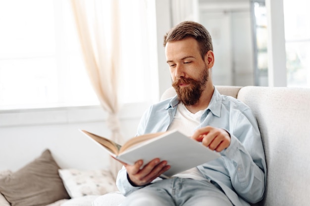 Joven hombre guapo con barba leyendo un libro en el sofá en casa