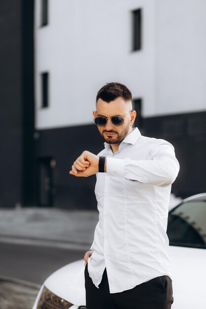 Joven hombre guapo con camiseta blanca y auriculares inalámbricos comprobando parámetros o hora en su reloj inteligente aislado sobre un fondo gris