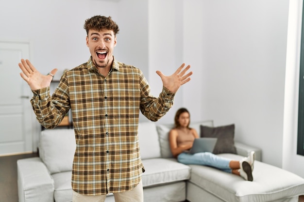 Joven hombre guapo en la sala de estar con su novia celebrando la victoria con una sonrisa feliz y una expresión de ganador con las manos levantadas