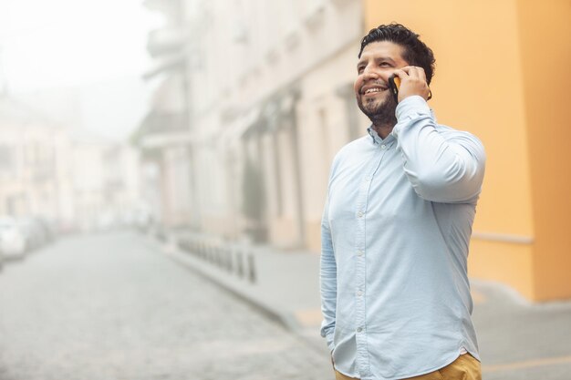 Foto joven hombre hispano alegre hablando por teléfono en una calle europea brumosa