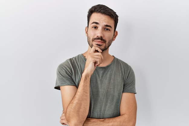 Joven hombre hispano con barba que usa una camiseta casual sobre un fondo blanco con la mano en la barbilla pensando en una pregunta expresión pensativa sonriendo con cara reflexiva concepto de duda