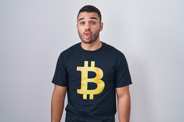 Joven hombre hispano con camiseta de bitcoin haciendo cara de pez con labios loco y cómico gesto expresión divertida