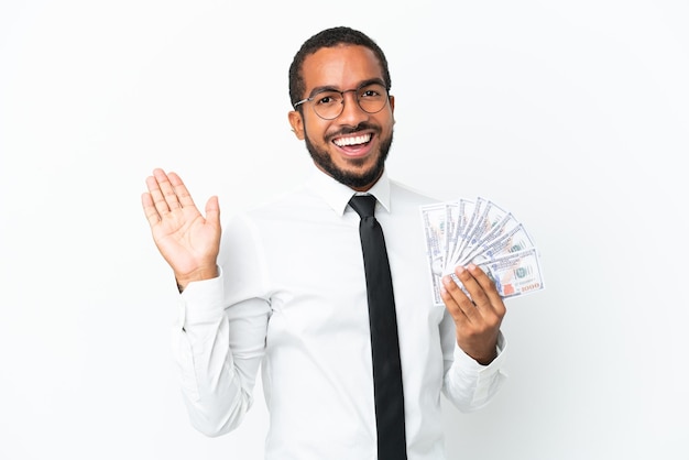 Joven hombre latino de negocios tomando mucho dinero aislado en fondo blanco saludando con la mano con expresión feliz