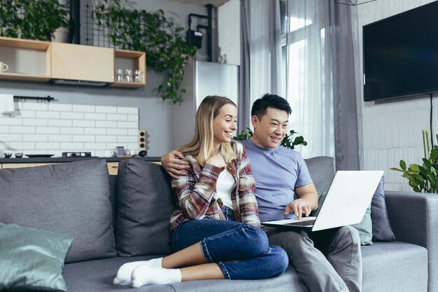 Joven hombre y mujer de familia divirtiéndose y felizmente sentados en casa usando una laptop y una pareja multirracial sonriente